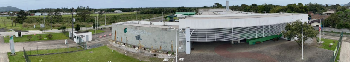 Imagem panorâmica do campus do Instituto Federal Catarinense, Campus São Francisco do Sul de 2024. O prédio principal possui uma fachada em pedra ardósia, com detalhes em brises brancos que cobrem parte do hall de entrada do campus, proporcionando uma entrada de luz indireta. À frente do prédio, há um caminho pavimentado e uma área aberta com gramado verde ao lado direito e também no lado esquerdo. No lado esquerdo da imagem, é possível ver a entrada do campus, com portões, guarita de segurança e uma placa com o logotipo do IFC. Ao fundo, uma área arborizada e montanhas são visíveis no horizonte.